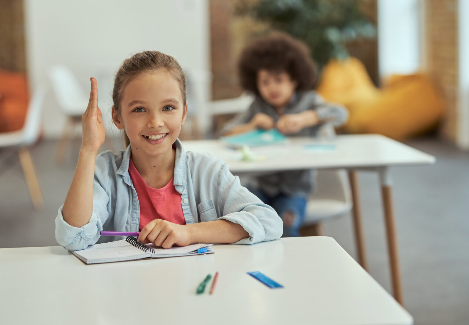 Active student. Cute little school girl smiling at camera raised her hand while sitting at the desk in classroom. Children and education concept